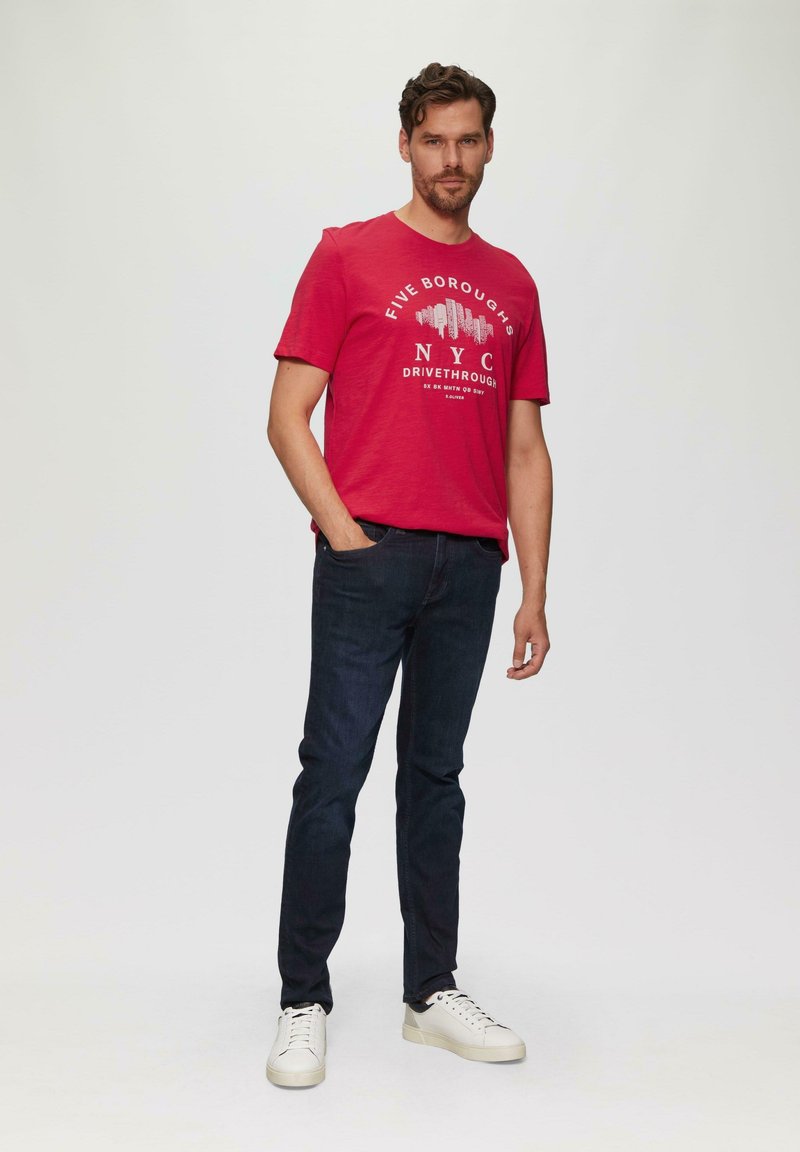 Rotes Baumwoll-T-Shirt mit dem Grafikdesign "FIVE BOROUGHS NYC DRIVETHROUGH", kombiniert mit dunklen Jeans und weißen Sneakers, steht vor einem neutralen Hintergrund.