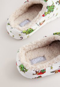 Textil-Hausschuhe mit einem weichen, beigen Innenfutter, verziert mit mehrfarbigen Grinch-Mustern auf weißem gestreiftem Hintergrund.