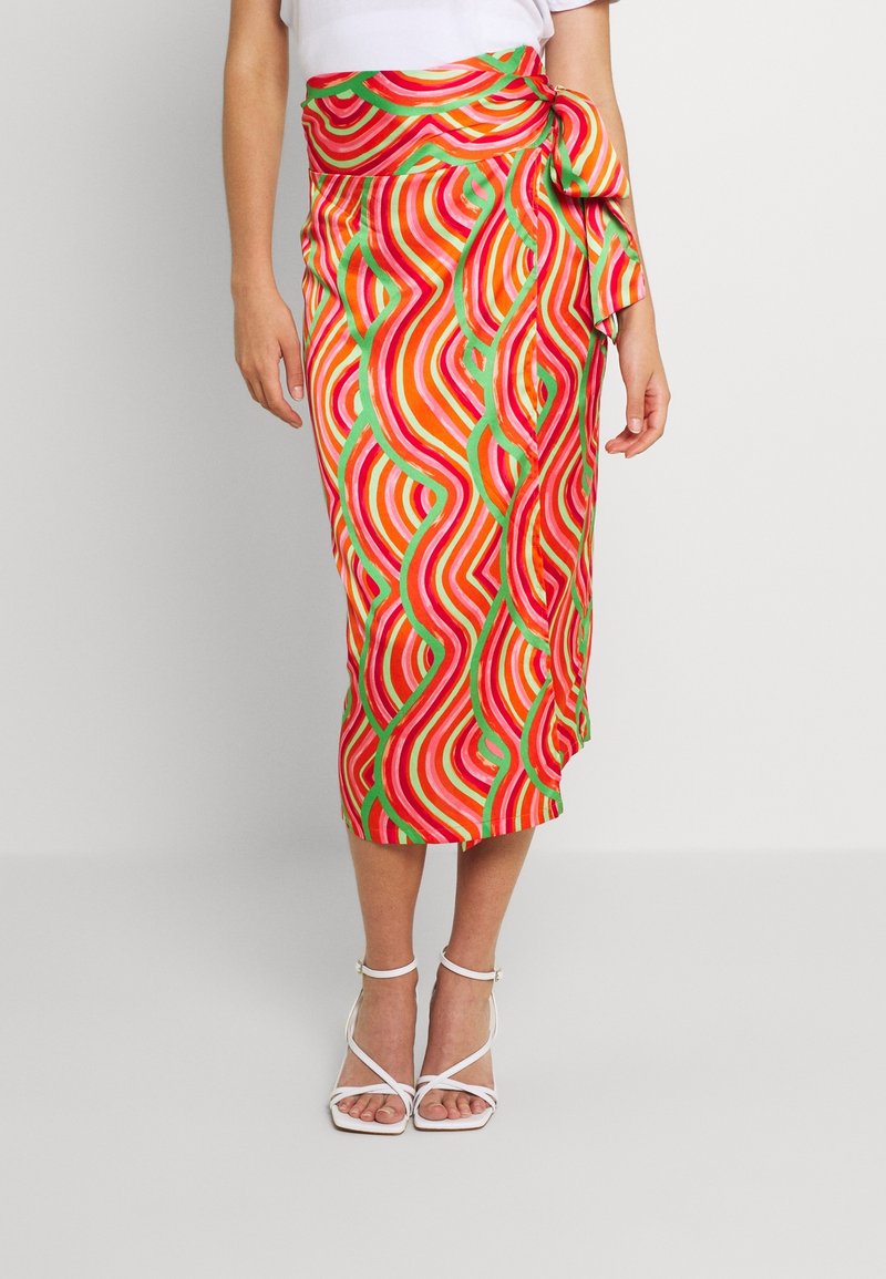 Femme portant une jupe midi colorée avec des rayures ondulées rouges, orange, roses et vertes et un nœud sur le côté, associée à des sandales blanches à talons.