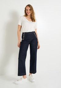 Jeans de tiro alto azul oscuro con botones de lado, diseño de pierna ancha, dobladillos acampanados, combinados con una camiseta blanca con escote en V y zapatillas blancas.