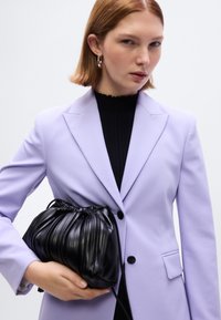 Μαύρη δερμάτινη τσάντα με κορδόνι και ριγωτή υφή, κρατημένη στο χέρι. Φορεμένη με λιλά σακάκι και μαύρο ζιβάγκο.