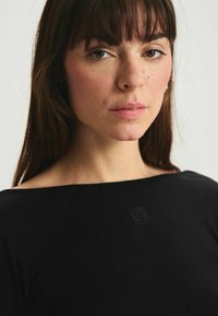 Haut noir avec un décolleté large, texture lisse et détail de logo discret sur la poitrine, encadré par de longs cheveux bruns et un maquillage neutre.