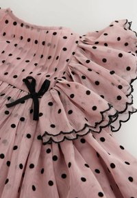 Tela rosa transparente con lunares negros, bordes fruncidos con ribete negro y un pequeño detalle de lazo negro.