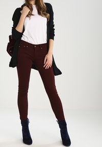 Burgundy smala jeans i kombination med en vit t-shirt och en svart kofta. Modellen bär blå anklarstövlar. Slät tyg med en figurnära passform.