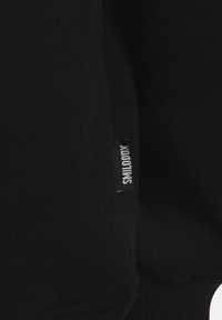 Primer plano de tela negra mostrando una pequeña etiqueta negra con el texto blanco "SMILODOX" cosida en la costura.