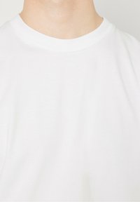 T-shirt en coton blanc avec un col rond, présentant une texture lisse. Le tissu a un léger éclat, mettant en valeur des détails de couture réguliers.