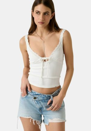 Donna che indossa una canottiera bianca con rifiniture in pizzo e pantaloncini di jeans azzurri strappati a vita alta, in posa con le mani vicino alle tasche.