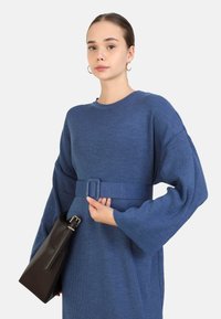 Blauses, geripptes Kleid mit langen Ärmeln und einem Gürtel-Detail an der Taille. Das Modell hält eine schwarze Handtasche mit einem metallischen Akzent.