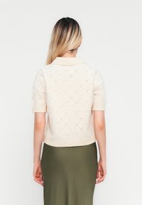 Žena s dlhými blond vlasmi má na sebe krátky rukáv béžový sveter s flitrami a olivovo zelenú sukňu, stojí čelom k bielej pozadi.