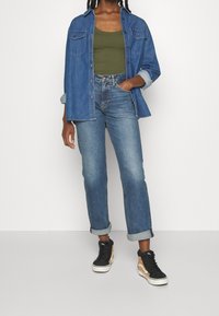 Camicia di jeans con due tasche sul petto, canotta verde oliva, jeans dritti azzurri chiari e sneaker alte nero-marroni, con le maniche arrotolate.