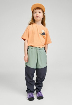 Małe dziecko w pomarańczowej czapce i koszulce, zielono-czarnych spodniach oraz fioletowych butach, stojące z jedną ręką w kieszeni na jednolitym tle.