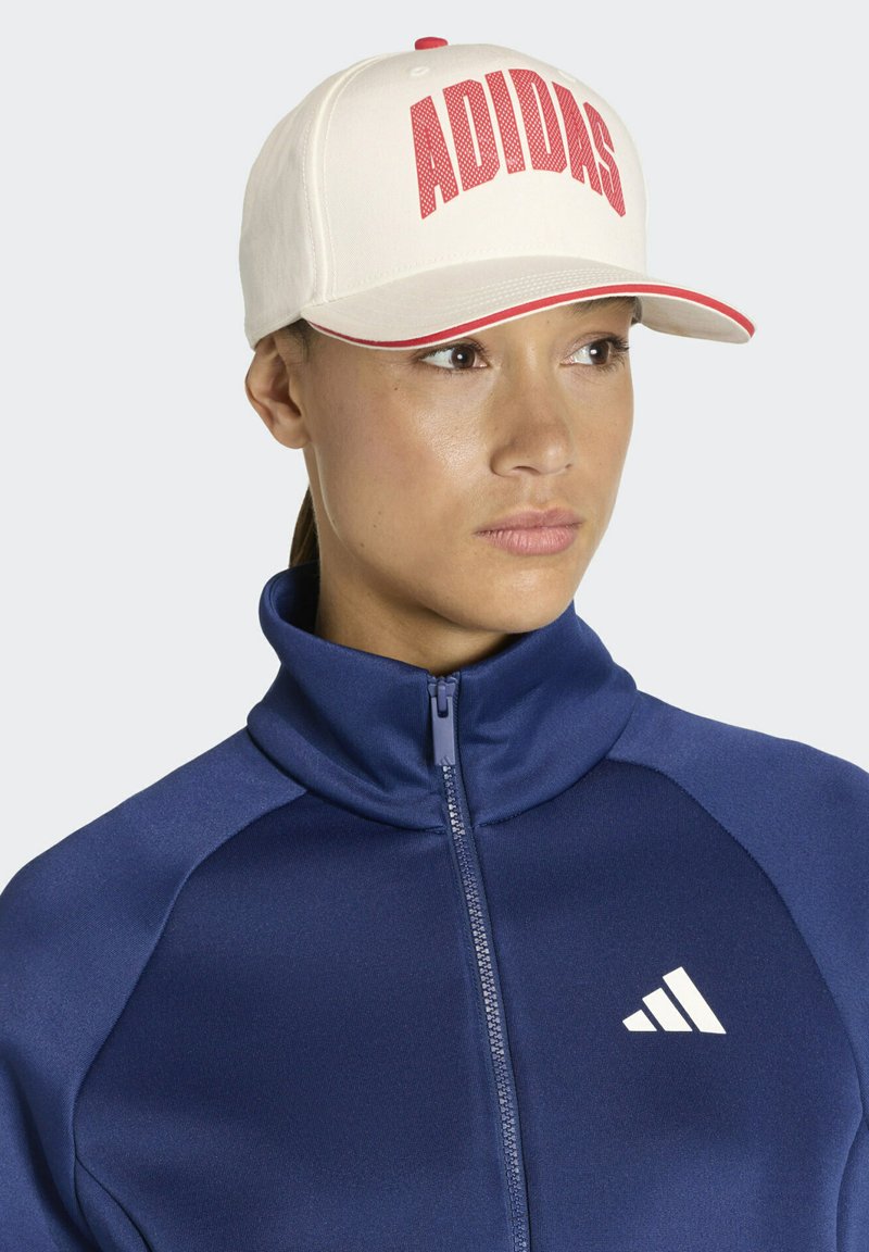 Gorra crema con el texto rojo "ADIDAS", con una visera curva y costuras contrastantes en rojo. Llevada con una chaqueta azul marino con cremallera.