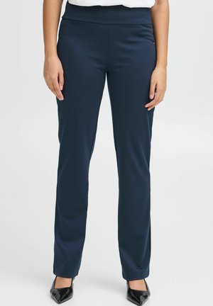 Navyblauwe broek van een gladde, elastische stof, met een brede tailleband en een recht pijpdesign, gecombineerd met zwarte puntige schoenen.