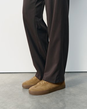 Personne portant un pantalon marron à jambes larges et des baskets en daim marron clair à lacets, debout sur un sol en béton gris.