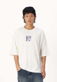 Hvid oversized t-shirt lavet af bomuld. Har et firkantet grafisk print centreret på brystet. Casual, boxy pasform med korte ærmer og rund hals.