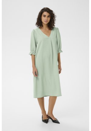 Ženska z kodrastimi lasmi, oblečena v svetlo zeleno midi obleko s puhastimi tridrskimi rokavi in črnimi sandali z ozkim koncem in z odprto peto.