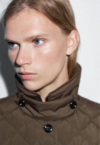 Gequiltede bruine jas met hoge kraag, voorzien van zwarte knopen en een gladde textuur. Het gezicht van het model is zichtbaar, met een nadruk op blauwe ogen en een natuurlijke huid.