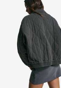 Ženska, ki se obrača na stran, nosi temno, prešito jakno z valovitimi vzorci in sivo krilo pred enobarvno belo podlago.