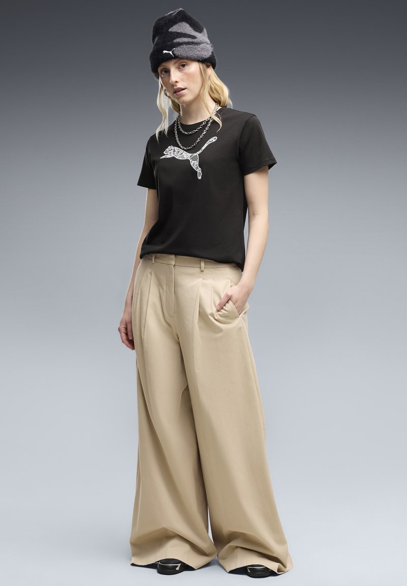 Νεαρή γυναίκα με μαύρο t-shirt Puma, μπεζ παντελόνι φαρδιάς γραμμής, μαύρο και γκρι σκουφάκι Puma και ασημένια στρωματοποιημένα κολιέ σε γκρι φόντο.