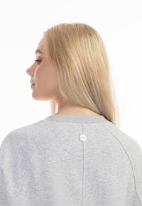 Ljusgrå sweatshirt med rund hals, raglanärmar och en vit knappdetalj på baksidan. Tyget verkar mjukt och texturerat.