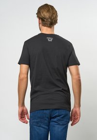 Sort bomulds t-shirt med korte ærmer, rund halsudskæring og et logo trykt i hvidt på den øverste del af ryggen. Båret sammen med blå denim jeans.