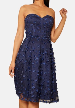Strapless navy blue midi dress with 3D floral appliques and fitted bodice, worn by a woman with one arm raised and the other down.