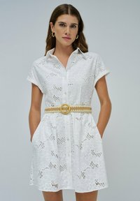 Mujer con un vestido corto de encaje floral blanco y mangas cortas, con botones en el frente y un cinturón tejido beige, de pie con las manos en los bolsillos.