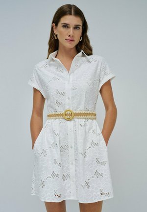 Femme portant une robe courte à manches courtes en dentelle florale blanche avec des boutons sur le devant et une ceinture tissée beige, se tenant avec les mains dans les poches.