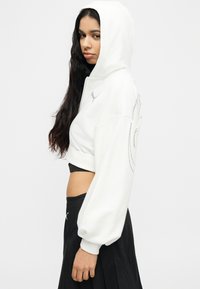 Γυναίκα με μακριά σκούρα μαλλιά, φορώντας λευκό κοντό φούτερ με κουκούλα ανασηκωμένη και μαύρη ψηλόμεση φούστα, γυρισμένη προφίλ σε λευκό φόντο.