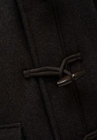 Zwart vilten bovenkleding met een toggle-sluiting en een gevlochten koord. De stof heeft een zachte textuur, met zichtbare stiksels en een zak.