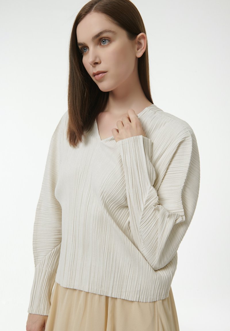 Cremfarbener gerippter Pullover mit V-Ausschnitt, langen Ärmeln und vertikaler Textur. Leichter Stoff mit lockerer Passform und cropped Länge.