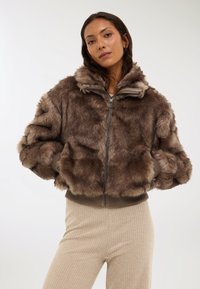 Giacca corta in finta pelliccia marrone con cappuccio, dotata di chiusura a zip, polsini a coste e una consistenza morbida. Abbinata a pantaloni beige a coste.