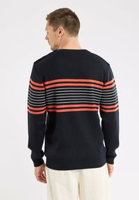 Homme de dos portant un pull noir avec des rayures horizontales rouges et blanches sur les épaules et les bras supérieurs, associé à un pantalon clair.