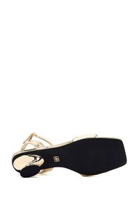 Gouden metallic hoge sandaal met een open achterkant, zwarte rubberen zool en dunne banden. Voorzien van een platte gesp voor aanpassing.