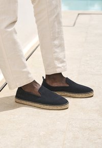 Espadrillas in camoscio nero con suola in juta intrecciata e pannelli elastici laterali, indossate con pantaloni di lino chiaro su una superficie testurizzata.