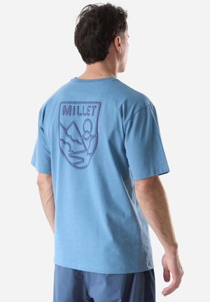 Mann trägt ein hellblaues T-Shirt mit dunkelblauem, gesticktem "MILLET"-Logo und Berggrafik auf dem Rücken, steht vor einem schlichten Hintergrund.