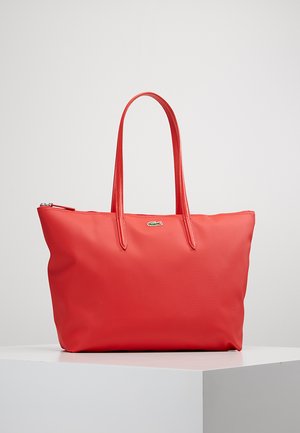Sac fourre-tout rouge en matériau lisse et durable, avec deux longues poignées, une fermeture éclair et un petit logo sur le devant.
