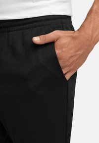 Sorte joggebukser med teksturert materiale, som har sidelommer og elastisk midje. Hånd hviler i lommen.