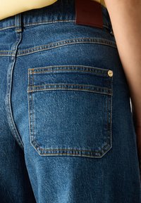 Jeans de ganga em azul médio com uma textura desbotada. Apresenta um único bolso traseiro com detalhe em metal dourado e um patch de couro com a marca.