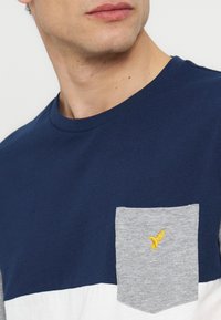 Άνδρας που φοράει μια μπλούζα σε μπλε ναυτικό και λευκό χρώμα με μια γκρι τσέπη που έχει μικρό κίτρινο λογότυπο πουλιού στο στήθος.