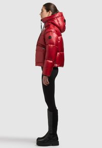 Sarkana pūku jaka ar kapuci, priekšpusē aizdares kabata un melns logotipa uzšuvums, kopā ar augstām melnām zābakiem, attēlota profilā uz neitrāla fona.