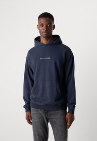 Hanorac albastru marin dintr-un material moale, cu un buzunar tip kangaroo și manșete ribbate. Logo-ul "Marc O'Polo DENIM" imprimat pe piept.