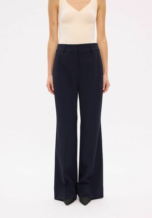 Femme portant un pantalon bleu marine taille haute à jambes larges, associé à un top beige sans manches, debout devant un fond uni.