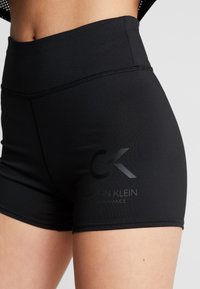 Nærbillede af en person iført sorte Calvin Klein Performance højtaljede træningsshorts med logo på venstre lår.