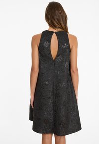 Femme vue de dos portant une robe noire sans manches avec un motif floral et une ouverture en forme de goutte d'eau à l'arrière supérieur.