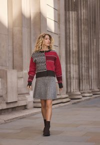 Värikäs neulepusero, jossa on punaisia ja mustia kuvioita, yhdistetty harmaaseen pliseerattuun hameeseen ja mustiin nilkkureihin, kävellen kivipäällysteisellä kadulla.