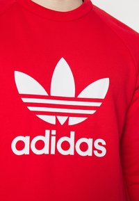 Röd sweatshirt i bomullsblandning, med en stor vit adidas-logotyp med trebladig design och horisontella ränder. Ribbad rund hals.