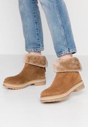 Brune ruskindsstøvler med foldede beige pelsmanchetter båret med lyseblå denimbukser på en hvid gulv.