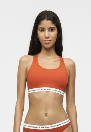 Femme portant un soutiens-gorge de sport orange et des sous-vêtements assortis avec des bandes élastiques blanches portant l’inscription Björn Borg, debout devant un fond uni clair.