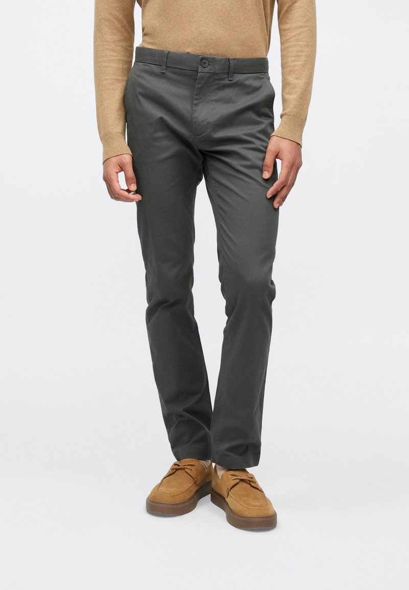 Pelēkas kokvilnas bikses ar šauru piegriezumu, ar pogu aizdari un sānu kabatām, kopā ar brūnām zamšāda kurpēm, uz vienkārša fona.